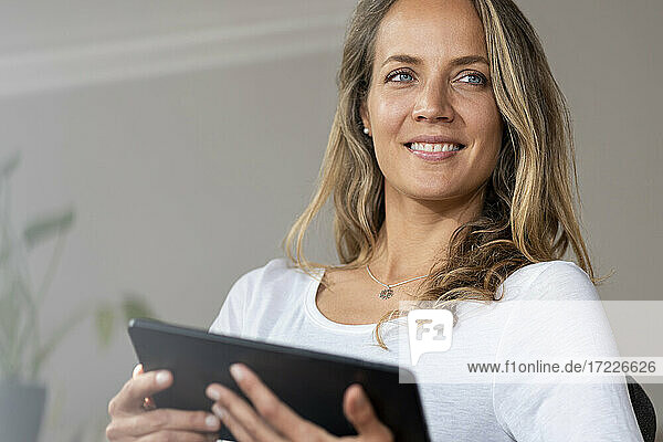 Lächelnde Frau mit blauen Augen  die wegschaut  während sie ein digitales Tablet zu Hause hält
