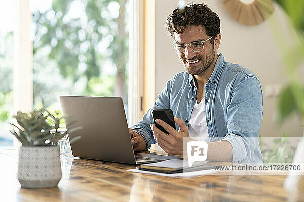 Lächelnder männlicher Freiberufler mit Brille  der ein Smartphone benutzt  während er mit einem Laptop im Büro zu Hause sitzt