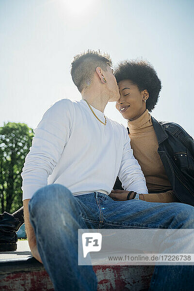Junger Mann küsst Freundin auf die Stirn  während er im Skateboardpark sitzt