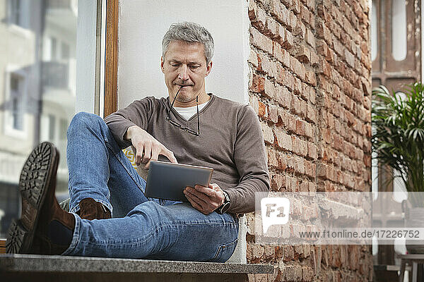 Man using digital tablet while holding eyeglasses in mouth at home
