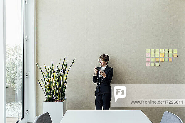 Lächelnde weibliche Fachkraft mit Mobiltelefon  die wegschaut  während sie im Büro an der Wand steht