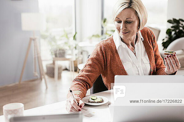 Lächelnde Geschäftsfrau  die in ihrem Büro zu Hause Brot isst und auf Papier schreibt