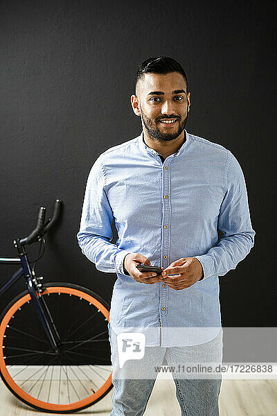 Lächelnder Mann mit Smartphone  der neben einem Fahrrad an einer Wand im Hintergrund steht
