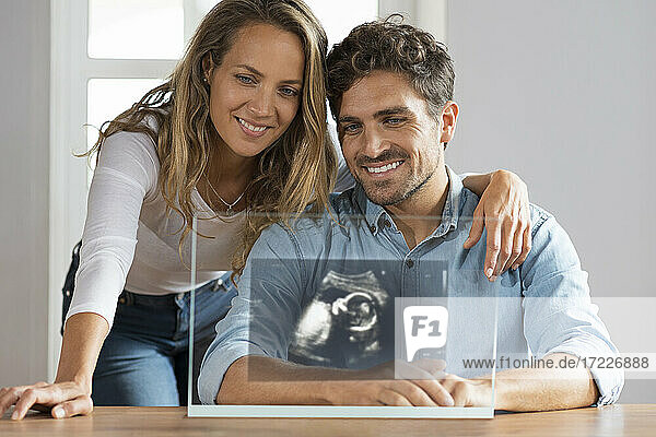 Glückliches Paar betrachtet Ultraschallbild auf transparentem Bildschirm zu Hause