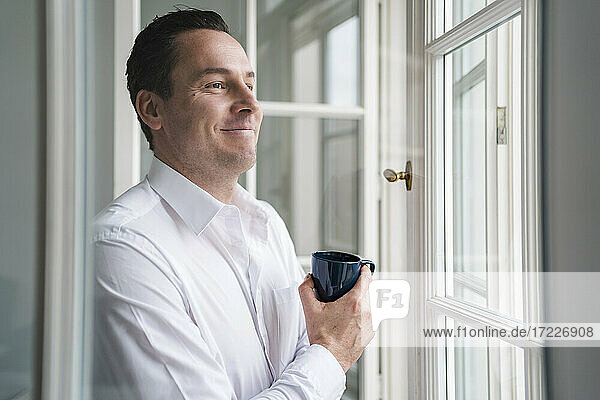 Männlicher Unternehmer  der eine Kaffeetasse hält und durch ein Fenster im Büro schaut