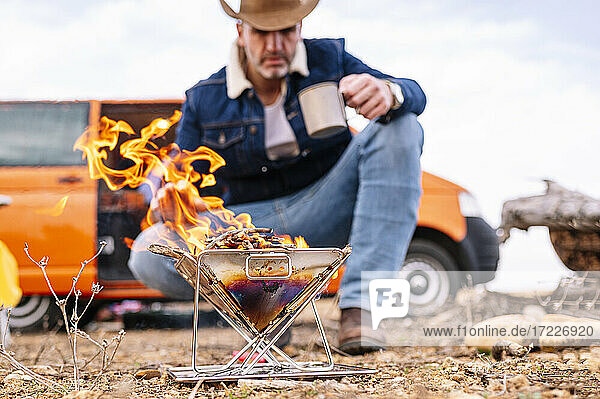 Mature man crouching near wood burning stove while camping on ground