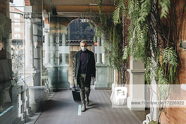 Mature man wearing protective face mask while walking with luggage at hotel corridor