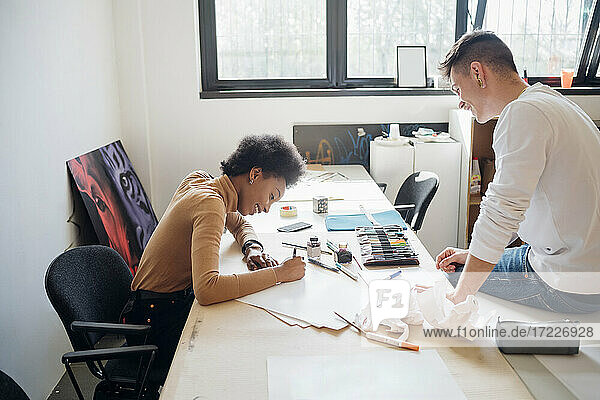 Lächelnder junger Mann  der seine Freundin beim Üben von Kalligraphie beobachtet  während er auf einem Tisch im Atelier sitzt