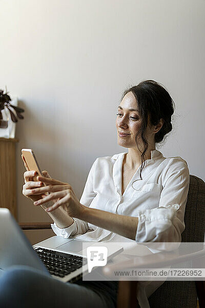 Lächelnde Frau  die ein Mobiltelefon benutzt  während sie mit einem Laptop auf einem Stuhl zu Hause sitzt