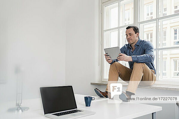 Männlicher Berufstätiger  der ein digitales Tablet benutzt  während er auf der Fensterbank im Büro sitzt