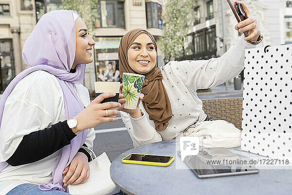 Freundinnen machen ein Selfie mit ihrem Smartphone und halten einen wiederverwendbaren Becher in einem Straßencafé in der Stadt.