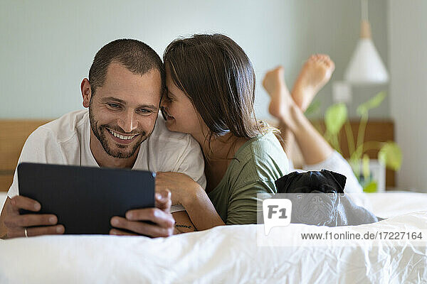 Frau umarmt Freund Blick auf digitale Tablette  während auf dem Bett im Schlafzimmer zu Hause liegen