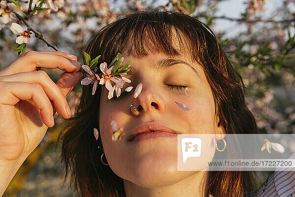 Smiling woman holding almond blossom branch by eyes during sunset