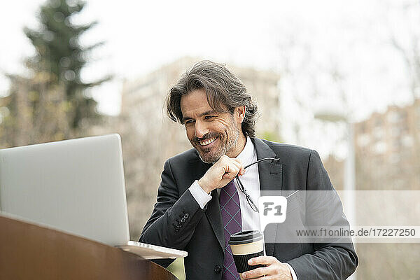 Glücklicher Geschäftsmann hält Einweg-Kaffeebecher und Brille und schaut auf einen Laptop