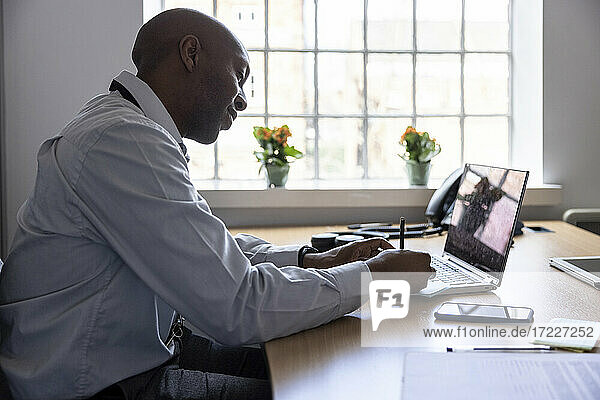 Männlicher Berufstätiger  der auf einem Laptop eine Haftnotiz schreibt  während er am Schreibtisch im Büro sitzt