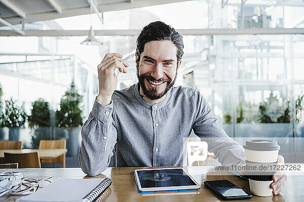 Lächelnder männlicher Freiberufler mit wiederverwendbarem Kaffeebecher durch drahtlose Technologien im Büro