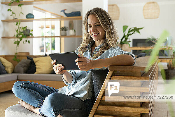 Lächelnde Frau  die ein Video über ein digitales Tablet ansieht  während sie auf der Couch im Wohnzimmer sitzt