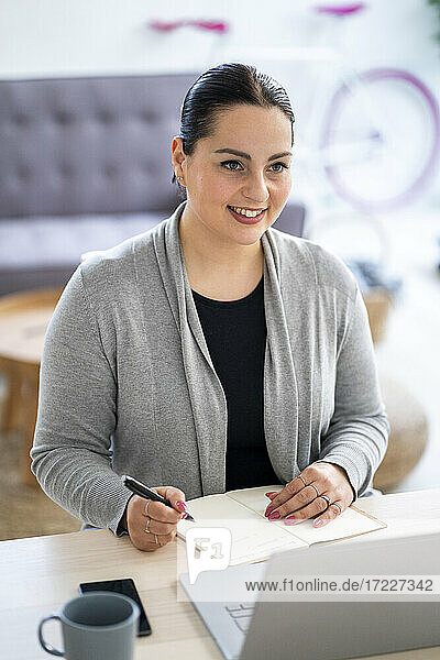 Lächelnde Geschäftsfrau  die einen Laptop benutzt und in ihr Tagebuch schreibt  während sie zu Hause arbeitet