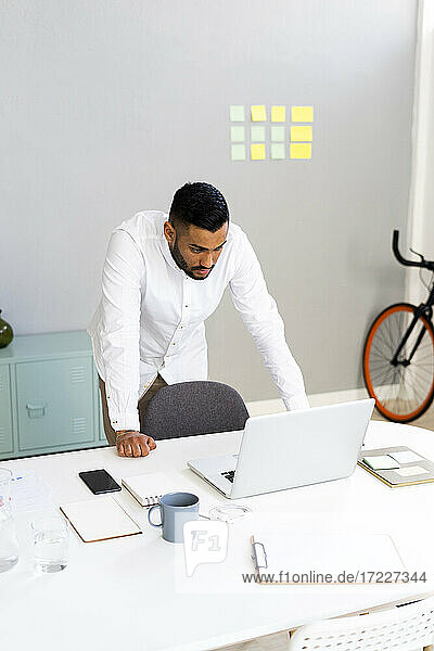 Geschäftsmann schaut auf einen Laptop und lehnt sich an einen Schreibtisch im Büro