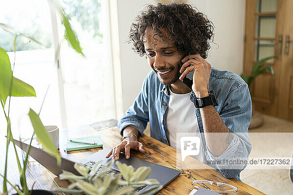 Männlicher Unternehmer  der mit seinem Smartphone telefoniert  während er zu Hause einen Laptop benutzt