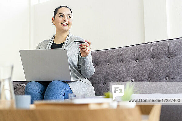 Lächelnde Frau  die wegschaut  während sie mit Kreditkarte auf einem Laptop online einkauft