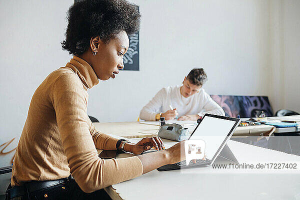 Junge Frau arbeitet am Laptop  während ein Mann am Tisch im Studio übt