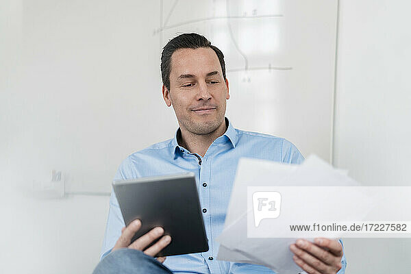 Männlicher Unternehmer  der ein digitales Tablet und Dokumente hält  während er im Büro sitzt