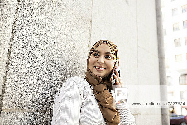 Junge Frau im Hidschab schaut weg  während sie mit einem Smartphone an der Wand telefoniert