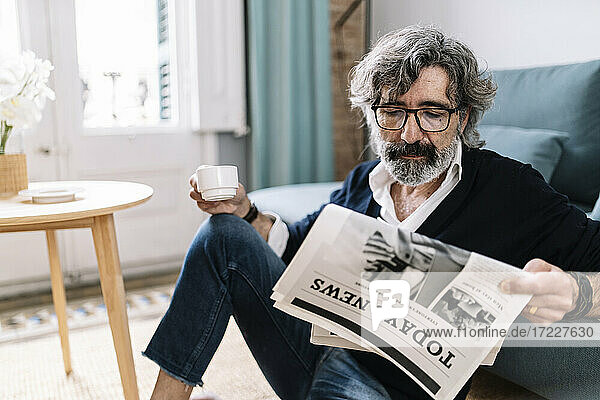 Hübscher Mann liest Zeitung und hält eine Kaffeetasse  während er zu Hause sitzt