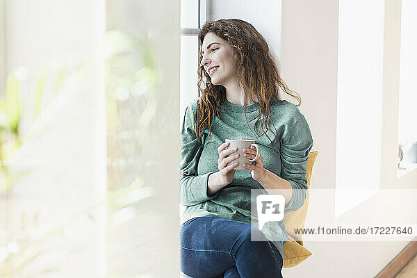 Lächelnde junge Frau mit Kaffeetasse  die nachdenklich am Fenster im Wohnzimmer sitzt