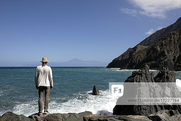 Älterer Mann schaut auf das Meer  während er auf einem Felsen vor dem Himmel steht
