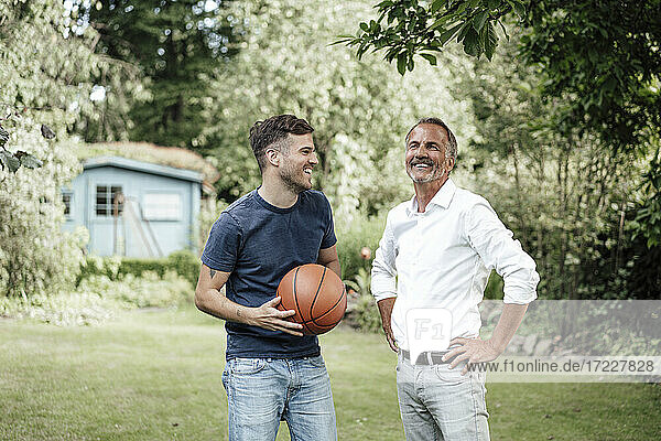 Lächelnder Vater mit Händen auf der Hüfte steht mit Sohn hält Basketball im Hinterhof