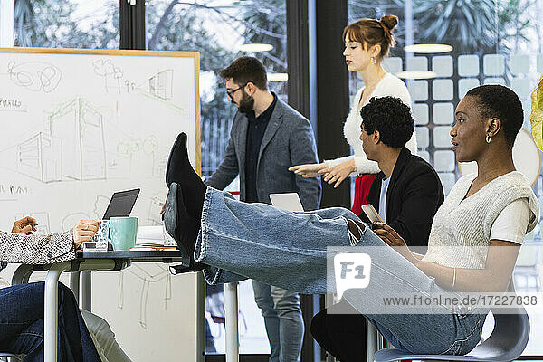 Geschäftsfrau mit Mobiltelefon lehnt Beine auf Tisch mit Kollegen im Hintergrund im Büro