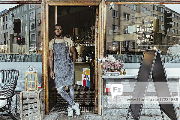 Smiling male owner leaning on doorway of delicatessen shop