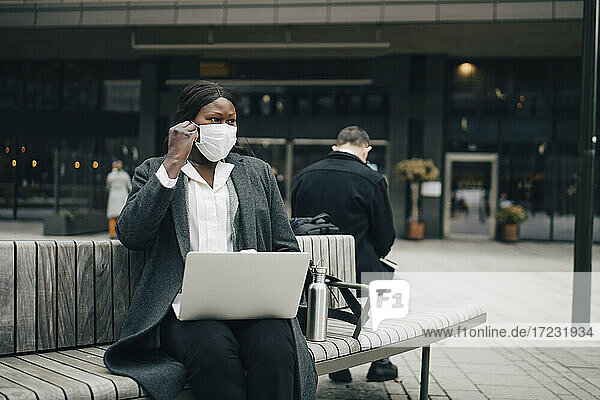Geschäftsfrau mit Laptop  die während der Pandemie auf einer Bank sitzt und wegschaut
