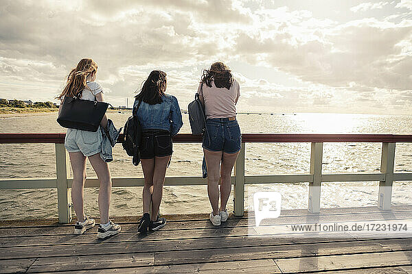 Rückansicht von Freundinnen  die sich bei Sonnenuntergang am Geländer des Piers anlehnen