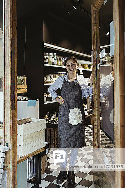 Portrait of smiling young female owner standing by door of delicatessen shop