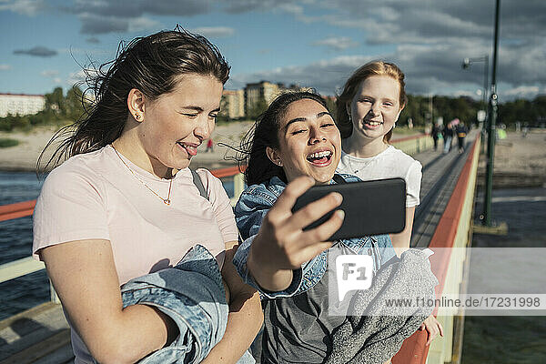 Fröhliche weibliche Freunde nehmen Selfie durch Smartphone auf Pier am Meer während sonnigen Tag