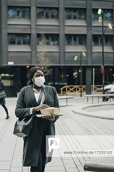 Geschäftsfrau mit verpackten Lebensmitteln und Laptop beim Spaziergang im Büropark während der Pandemie