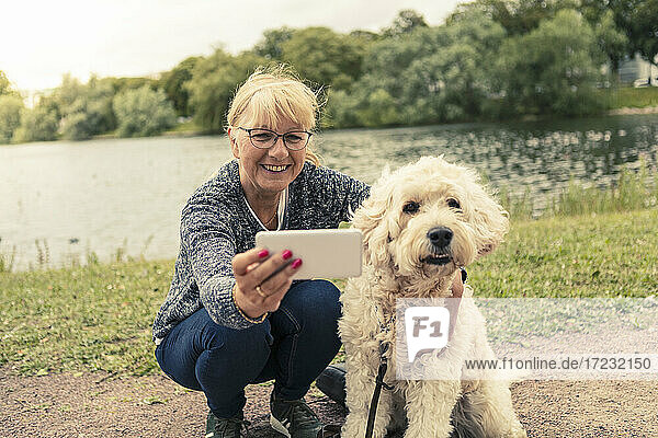 Lächelnde Frau nimmt Selfie mit Hund durch Smartphone am Seeufer