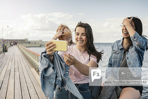 Glückliche weibliche Freunde nehmen Selfie durch Smartphone auf Pier gegen Meer