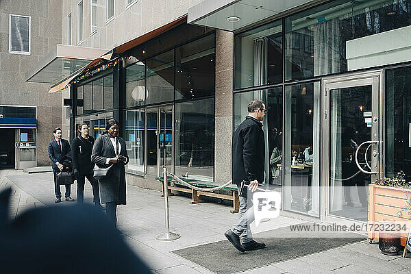 Geschäftsmann  der in ein Geschäft geht  während männliche und weibliche Kollegen auf dem Fußweg in einer Reihe stehen  während COVID-19