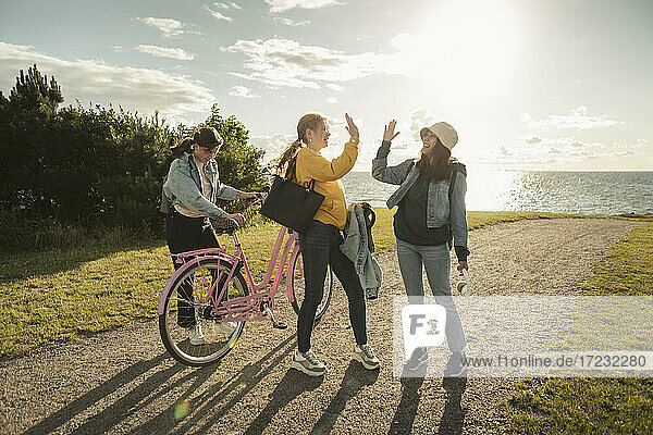 Female friends doing high-five while teenage girl with bicycle at lakeshore on sunny day