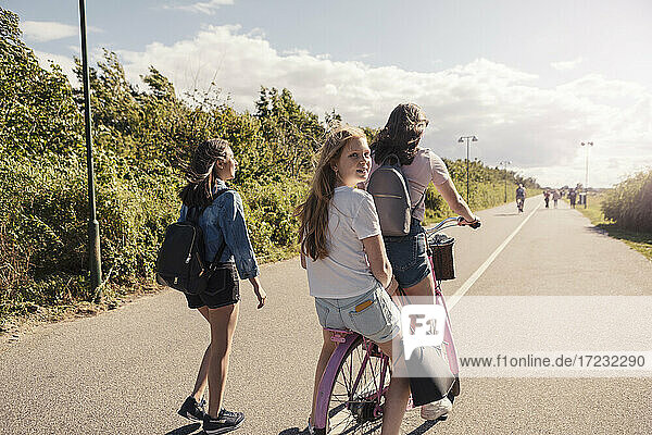 Female friends cycling while teenage girl walking on road during sunny day against sky