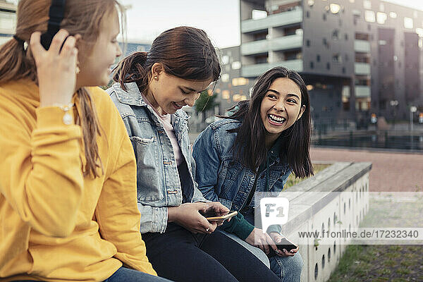 Happy female friends using smart phone while talking with each other at retaining wall
