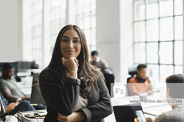Lächelnde Unternehmerin mit Hand am Kinn im kreativen Büro