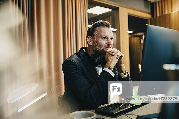 Lächelnde männliche Unternehmer mit Hand auf Kinn arbeiten über Laptop im Büro