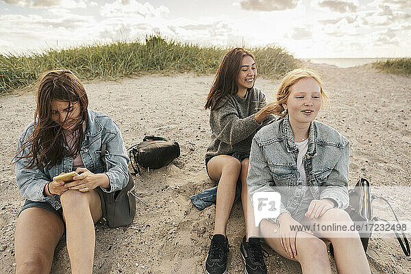 Teenage girl using smart phone while female friends sitting on sand at beach