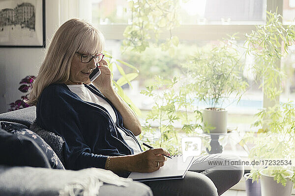 Reife Frau spricht auf Smartphone  während mit Buch zu Hause sitzen