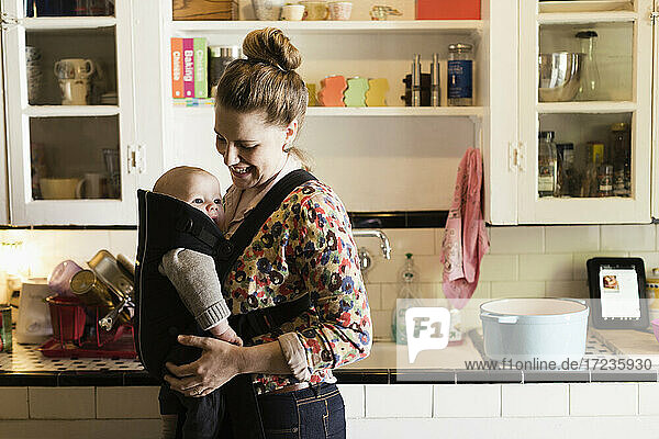 Mid adult mother with baby son in sling in kitchen